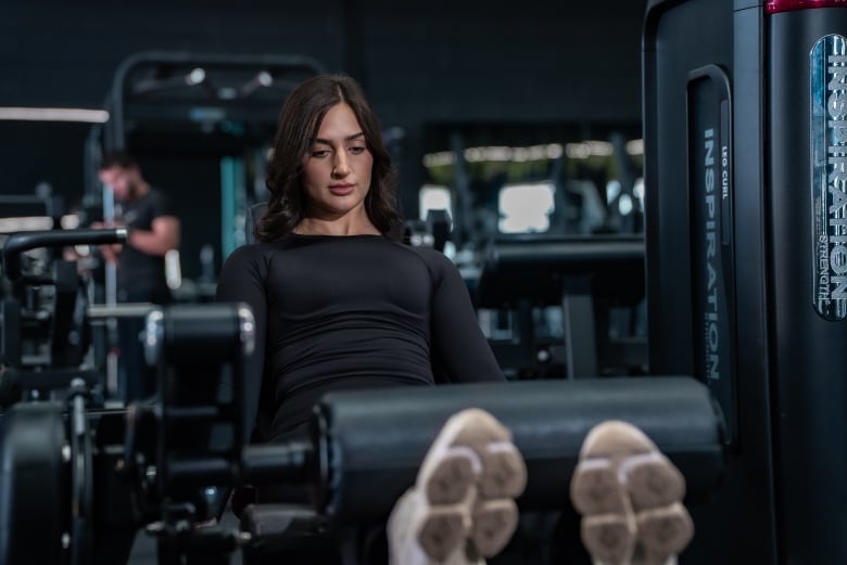 A young dark-haired woman uses a leg exercise machine in the gym. She is wearing a nice black workout outfit. 