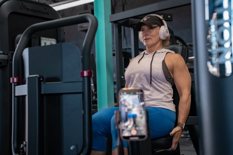 A woman in a baseball cap sits on an abductor exercise machine. She is wearing a white workout tank and blue leggings. There is a tripod with a phone camera recording him in front of him. 