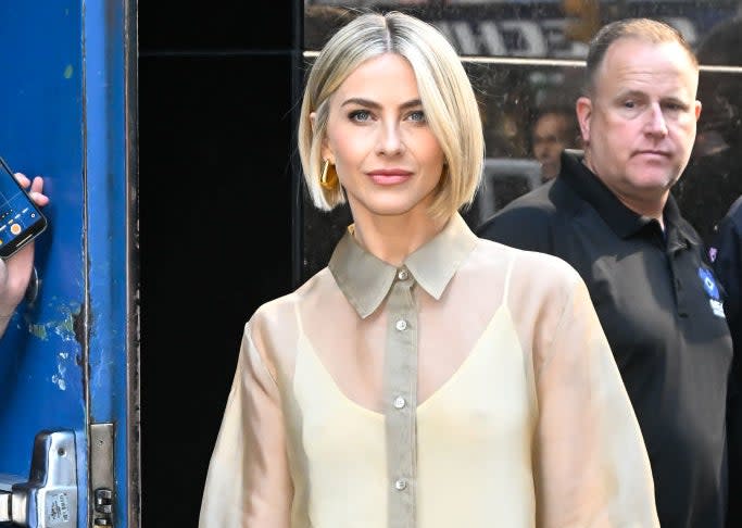 Julianne Hough, wearing a thin, long-sleeved blouse and tight dress, holding a white clutch, stands outside the building with a man behind.