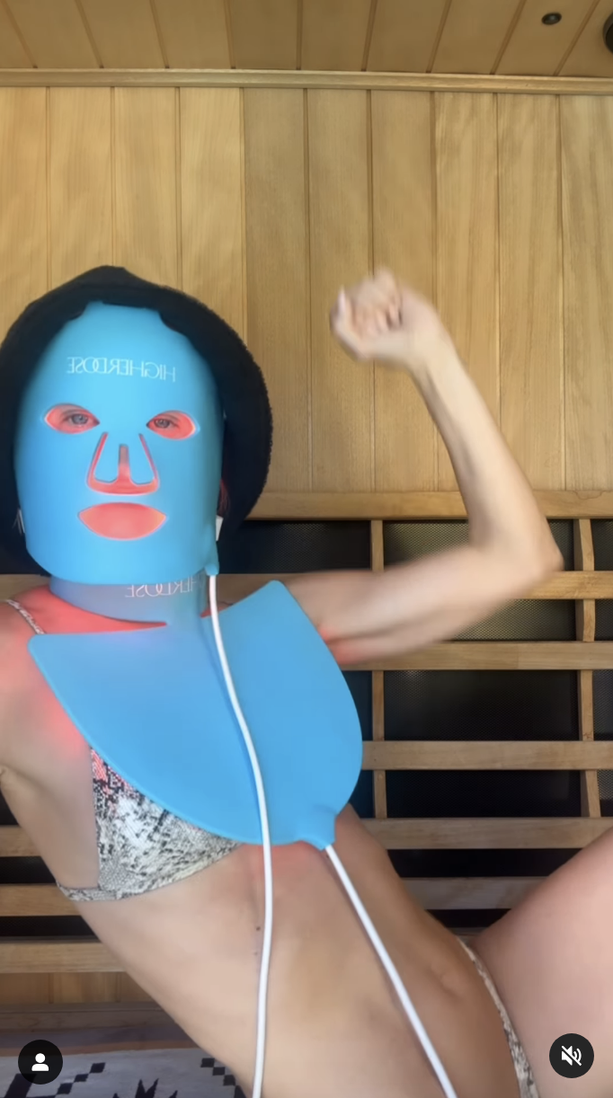 A person wearing a turquoise LED face mask and a matching neck piece, poses awkwardly in a bikini, in a wooden sauna-like room.